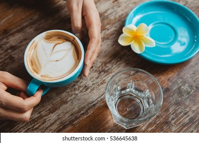 Young woman holding a cup of morning coffee cappuccino served on wooden table with plate, frangipani flower and a glass of water