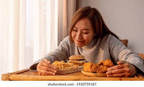 A Young Woman Holding And Eating A Hamburger, French Fries And Fried Chicken On The Table At Home