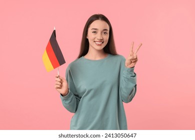 Young woman holding flag of Germany and showing V-sign on pink background