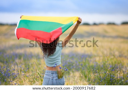 Young woman holding flag of Lithuania in a rye field with blue cornflowers. Back view. Lithuanian Flag Day. Independence restoration Day. Travel, learn lithuanian language and love Lithuania concept. 