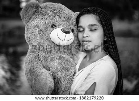 Young woman holding a giant fluffy teddy bear inside a blue river in a wild natural scenery. The girl seems nostalgic and thinking about her past.