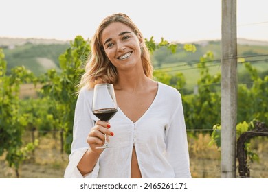 Young woman holding glass of red wine inside vineyard during sunset time while smiling on camera - Powered by Shutterstock