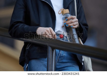 Young woman holding glass water bottle and lollipop candy in hand. Unrecognizable female person wearing black leather jacket and classic blue jeans on a walk