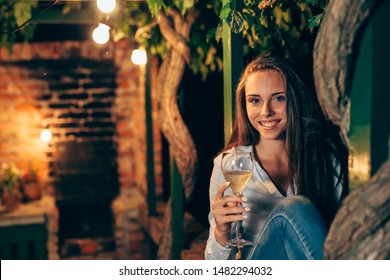 Young Woman Holding Glass Of Wine Sitting At Home In Backyard. Night Scene