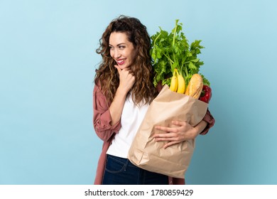 Young Woman Holding A Grocery Shopping Bag Isolated On Blue Background Looking To The Side And Smiling