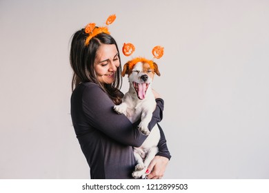 Young Woman Holding Her Cute Small Dog Over White Background. Matching Pumpkin Diadems. Halloween Concept. Indoors