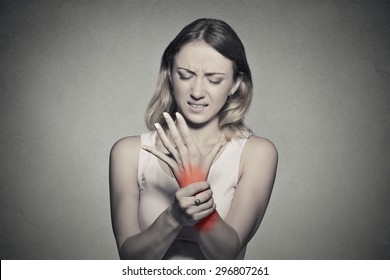 Young woman holding her painful wrist isolated on gray wall background. Sprain pain location indicated by red spot. Negative face expression 
