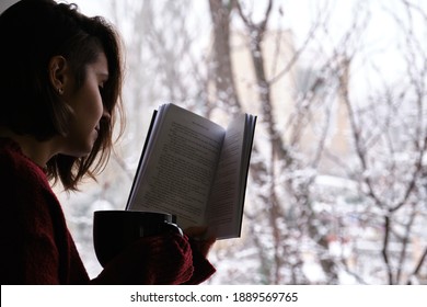 Young Woman Holding A Hot Coffee Or Tea Cup, Reading A Book By The Window In A Snowy Winter Day. Cozy At Home Concept.