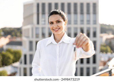 Young woman holding house keys with smile standing on urban rooftop. Real estate agent or homeowner concept, cityscape background. Happy feelings, achievement, success in property ownership. - Powered by Shutterstock