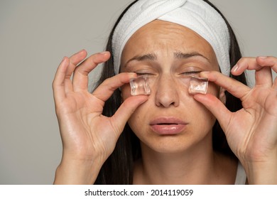 a young woman holding ice cubes under her eyes on a gray background