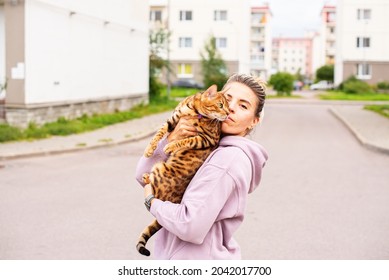 Young Woman Holding And Loving Her Bengal Cat Outside. Walk With Pet, Concept Of Pet Care, Cat On Harness, Daily Walking, Jogging, Training And Any Outdoor Recreations With Lovely Pet. 