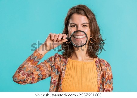 Young woman holding magnifier glass loupe on healthy white teeth, looking at camera with happy expression, showing funny silly face smiling mouth. Brunette girl isolated on blue studio background