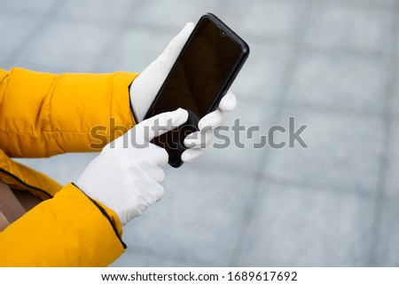 Young woman holding a mobile phone in her hands wearing latex protective gloves at the street in public place. Coronavirus spreading precautions measures concept.