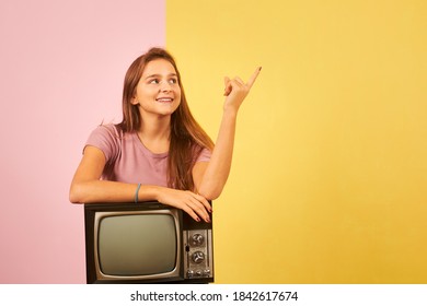 Young Woman Holding Old Retro Tv Sitting Against Yellow And Pink Background Pointing With Finger To The Side