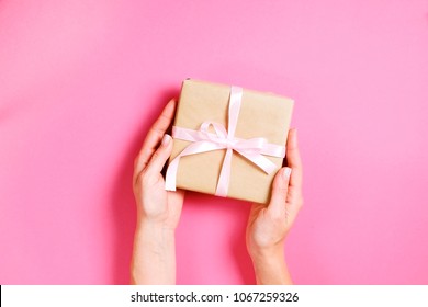 Young woman holding one single present wrapped in hand made blank craft paper gift wrap, satin bow. Female hands, simple giftbox, wrapping, giftwrap, silk ribbon. Pink background, close up, copy space