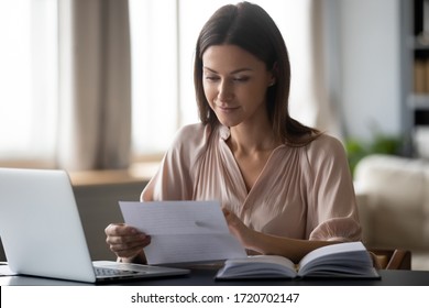 Young Woman Holding Paper, Reading Good News In Letter, Sitting At Desk With Laptop At Home, Beautiful Female Received Correspondence, Bank Statement Or Notification, Pleasant Message