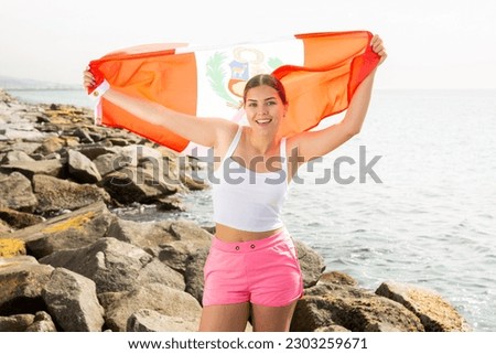 Young woman holding Peru waving flag at coastline against of sunny bright sea