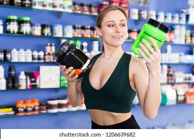 Young Woman Holding Plastic Jar With Sport Nutrition, Reading Label Carefully Before Buying In Shop