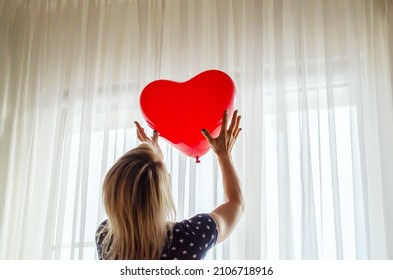 Young woman holding red heart shaped baloon against the window at home. Celebrating valentines day, wedding and love day, women's day, World Heart Day, birthday. Silhouetted against the sun indoor