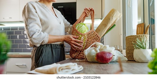 Young woman holding reusable mesh bag with fruits, vegetables and bread in the kitchen. Zero waste and sustainable packaging concept. Horizontal panoramic banner. - Powered by Shutterstock