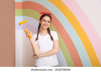 Young Woman Holding Roller Near Wall With Painted Rainbow Indoors