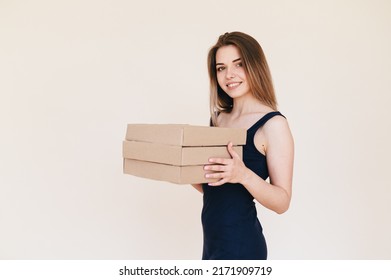 Young Woman Holding Stack of Cardboard Boxes on Beige Background. Girl with a Smile on her Face in a Black Dress Received a Delivery. Food delivery