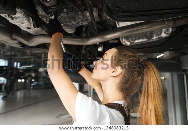 Young\
woman holding tool, fixing car. Girl working undercarriage of\
automobile, auto lifted on bridge. Female mechanic in black gloves\
looking up, repairing vehicle in service\
station.