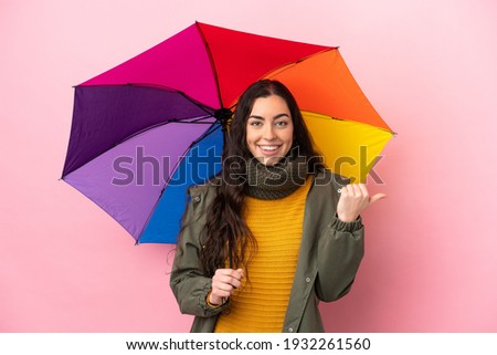 Young woman holding an umbrella isolated on pink background pointing to the side to present a product
