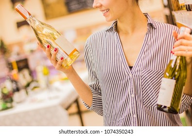 Young Woman Holding Wine Bottles In Hands And Smiling While Choosing Wine In Grocery Shop. Beautiful Girl Doing Shopping In Big Supermarket. Woman Looking At Blank Label With Copy Space On Wine Bottle