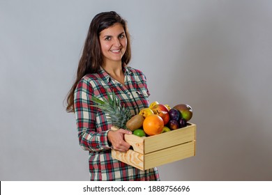 Young Woman Holding Wooden Box Full Of Fresh Fruit. Smiling Brunette With Wood Crate Filled With Juicy Healthy Food In Studio. Happy Girl In Flannel Shirt Showing Grapefruit, Plums, Mango And Others.