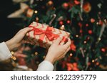 Young woman holding wrapped Christmas present with a red ribbon, standing near Christmas tree.