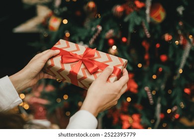 Young woman holding wrapped Christmas present with a red ribbon, standing near Christmas tree. - Powered by Shutterstock