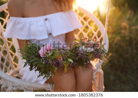 Young woman holding wreath made of beautiful flowers on swing chair outdoors at sunset, closeup