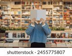 A young woman holds a book in front of her face cover, standing amidst a vibrant and varied bookstore. Shelves of books create a cozy, intellectual backdrop.