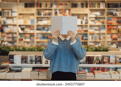 A young woman holds a book in front of her face cover, standing amidst a vibrant and varied bookstore. Shelves of books create a cozy, intellectual backdrop. - Powered by Shutterstock