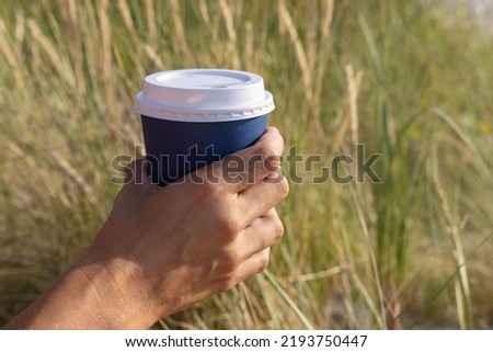 Young woman holds a disposable cup of coffee on on a sunny morning at the beach. Take away coffee at the sea. 