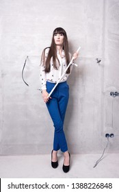 Young Woman Holds A Folding Meterstick Carpenter's Ruler Diagonally In Front Of Her While Standing In Front A Gray Wall Of An Apartment Undergoing Renovation