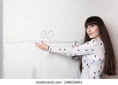 Young Woman Holds A Folding Meterstick Carpenter's Ruler In Front Of A Measurement On A Gray Wall Of An Apartment Undergoing Renovation