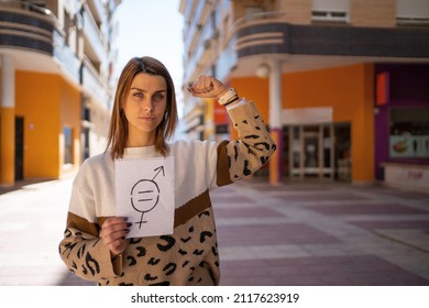 Young Woman Holds A Gender Equality Sign While Raising Her Arm Showing Her Strength In The Middle Of The Street During A Sunny Day.