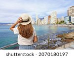 Young woman holds hat on promenade in front of the skyline from Benidorm Downtown, Spain