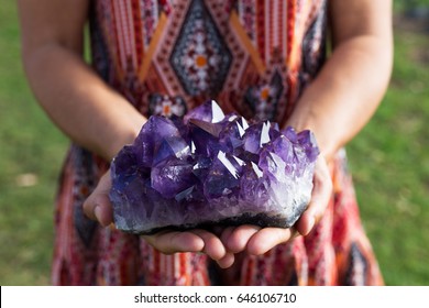 A young woman holds a large cluster of amethyst crystals as they shimmer in the sunlight.