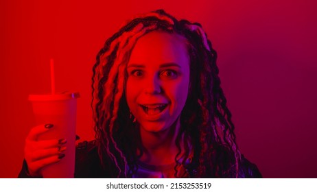 Young woman holds large cup of soft drink with straw in dark room. Portrait of female posing with nonalcoholic beverage in darkness illumination of red light - Shutterstock ID 2153253519