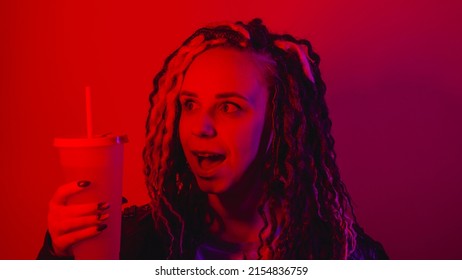 Young woman holds large cup of soft drink with straw in dark room. Portrait of female posing with nonalcoholic beverage in darkness illumination of red light - Shutterstock ID 2154836759