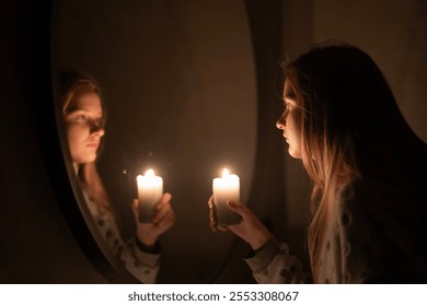 A young woman holds a lit candle, gazing at her reflection in a mirror. The warm glow of the candle softly illuminates her features, creating a serene and introspective ambiance filled with calm and - Powered by Shutterstock