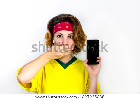 A young woman holds a lodon on her lips, thereby to close,  her mouth with her hand, a secret. A young woman is holding her mobile phone and smartphone on an isolated background. Copy space.