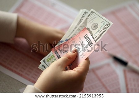 A young woman holds the lottery ticket with complete row of numbers and dollar bills on the lottery blank sheets background. Lottery winner concept