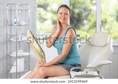 Young woman holds mirror in hands and admires reflection. Smoothing wrinkles, smoothing skin tone, therapeutic and cosmetic procedures in cosmetology salon