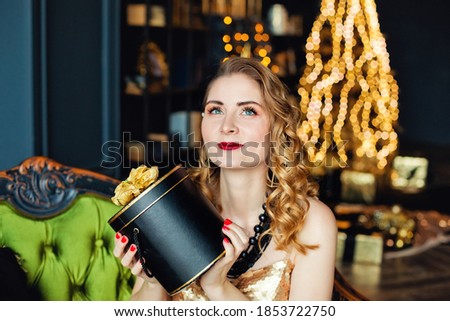 A young woman holds a string of beads against the background of a Christmas tree with gold ornaments. Stylish women's, fashionable evening wear and accessories, makeup. Happy New Year.