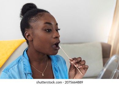 Young Woman Holds A Swab Into Her Mouth For The Coronavirus COVID-19 Home Test. Woman Doing DNA Test With Cotton Swab At Home. Test For Home Use. African Young Girl Using A Home Test Kit For COVID-19.