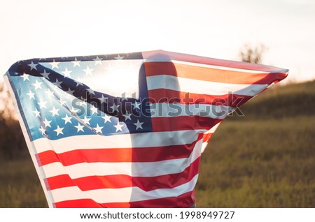 A young woman holds the US national flag in a field at sunset. 4th of July, Independence day. American freedom concept. Selective focus